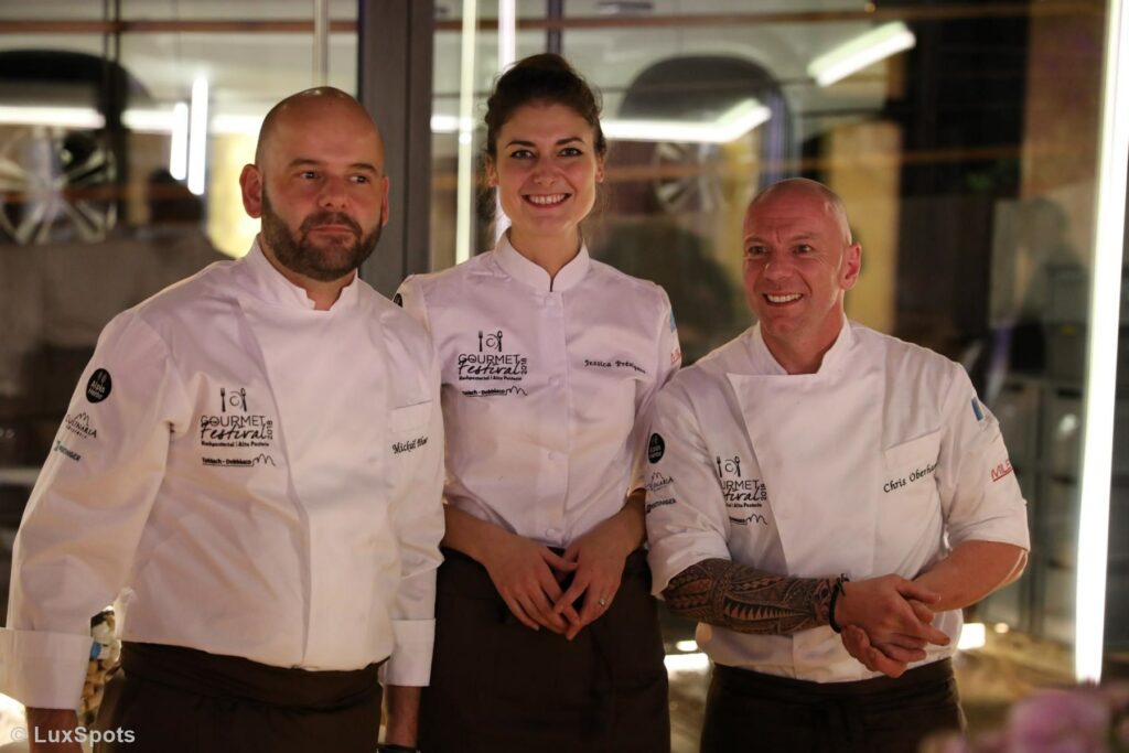 Drei Köche in weißen Jacken stehen lächelnd zusammen bei einer gemeinsamen Veranstaltung.