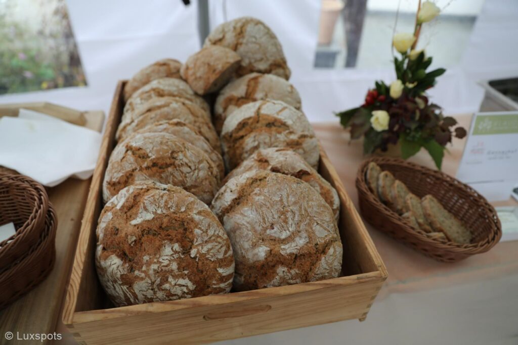 Ein Holztablett gefüllt mit runden, rustikalen Brotlaiben auf einem Tisch, daneben ein kleiner Korb mit geschnittenem Brot und eine Vase mit Blumen.