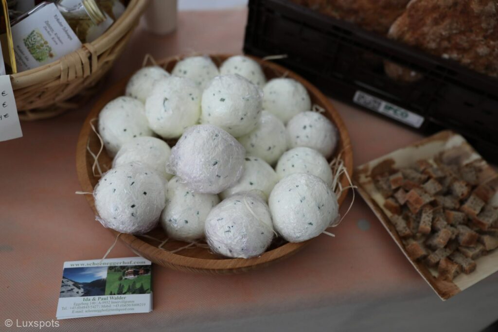 An einem Marktstand steht eine Holzschale mit runden Käsesorten. Daneben liegen Visitenkarten und ein Tablett mit Brotproben.