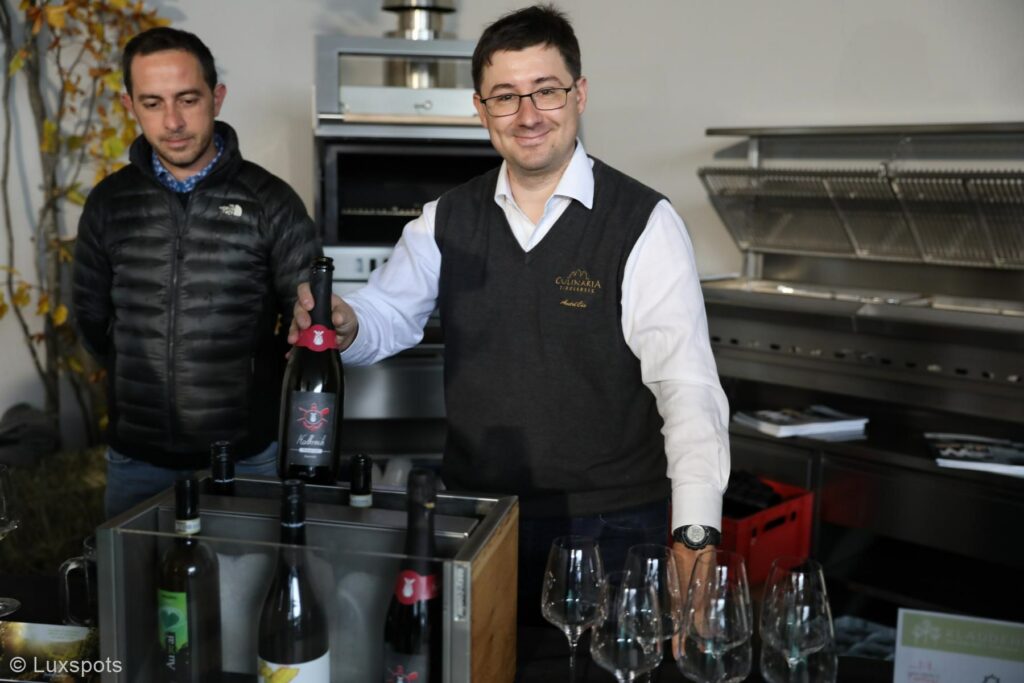 Zwei Männer stehen bei einer Weinauslage, ein Mann hält eine Weinflasche und auf dem Tisch stehen verschiedene Weingläser.