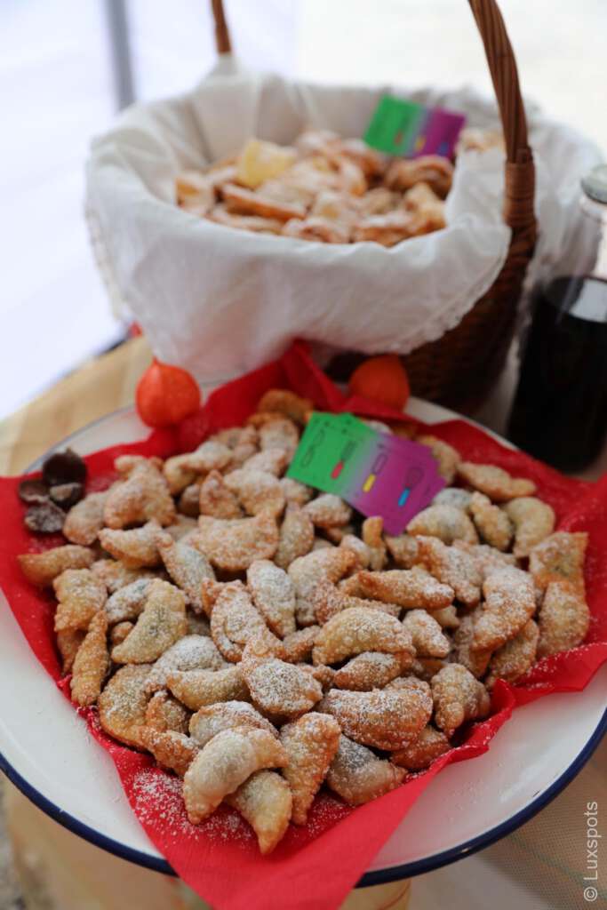 Ein Teller und ein Korb gefüllt mit halbmondförmigem Gebäck, bestäubt mit Puderzucker, auf einem Tisch stehend, daneben eine Flasche und bunte Dekorationen.