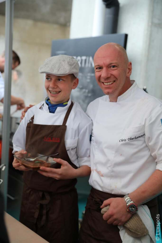 Zwei lächelnde Köche in weißen Uniformen und braunen Schürzen stehen zusammen; einer hält ein Paket, während der andere eine Schiebermütze trägt.