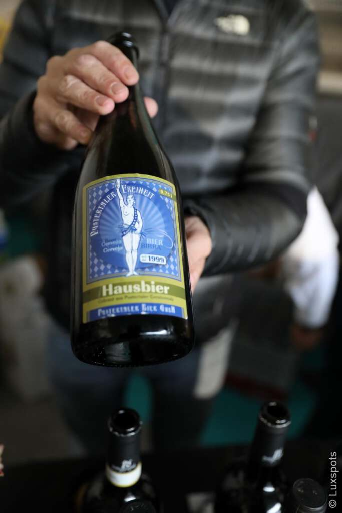 Eine Person hält eine Flasche Hausbier mit einem blau-goldenen Etikett. Mehrere andere Flaschen stehen darunter auf dem Tisch.