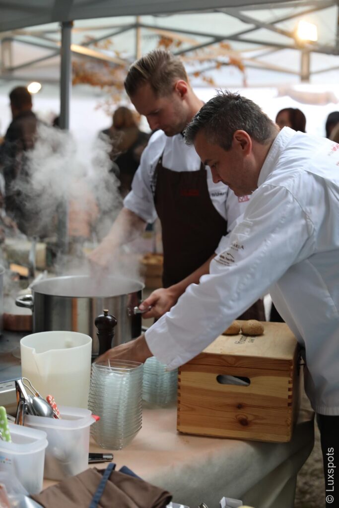 Zwei Köche in weißen Uniformen bereiten an einer dampfenden Kochstation im Freien, umgeben von Küchengeräten und Zutaten, Essen zu.
