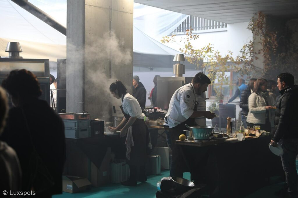 Unter fast freiem Himmel bereiten Köche Essen zu. Umgeben von Kochgeräten und Zutaten. Rauch steigt aus einem Grill auf und Menschen versammeln sich an den Kochstationen.