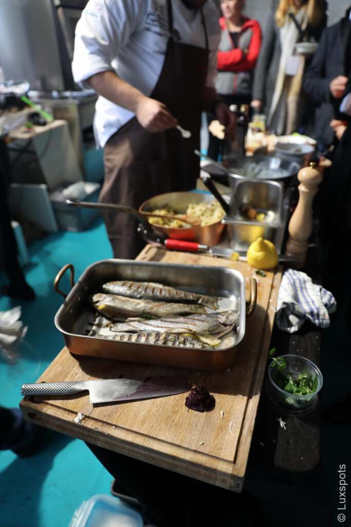 Koch bereitet in einer Küche gegrillten Fisch zu. Mehrere Gerichte, darunter in Folie eingewickelter Fisch, Kräuter und eine Zitrone, stehen auf einer Holztheke. Leute stehen daneben und schauen zu.
