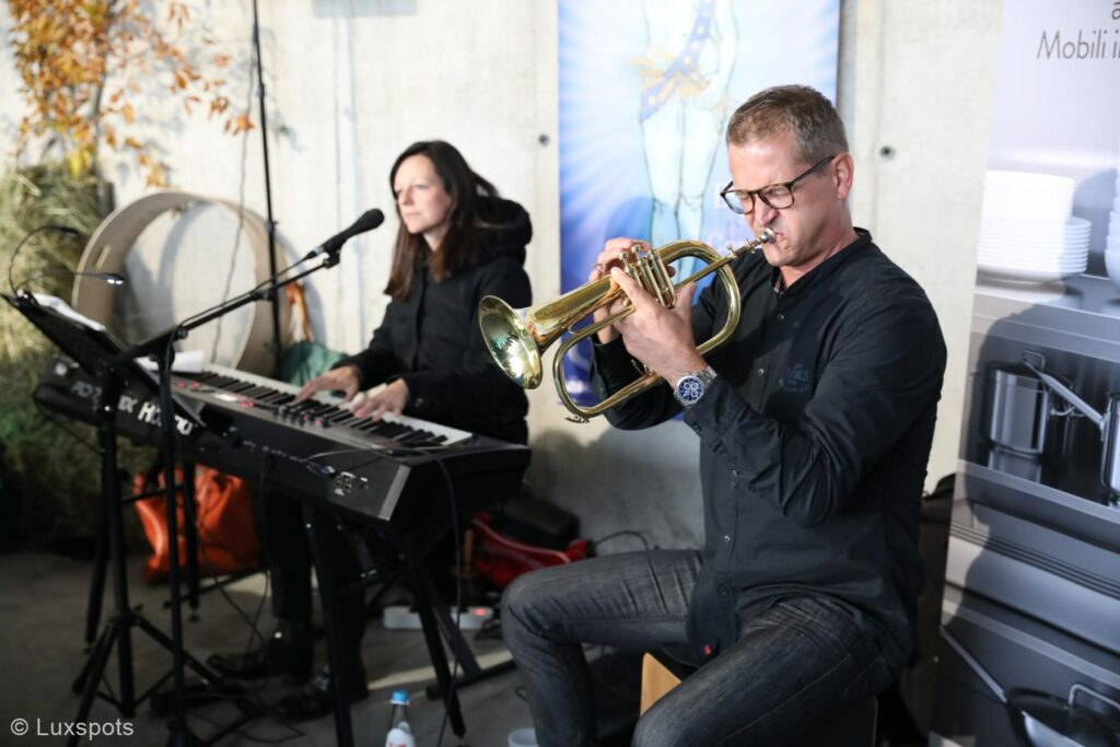 Ein Mann spielt Trompete, während eine Frau in einer lockeren Aufführungssituation Keyboard spielt.
