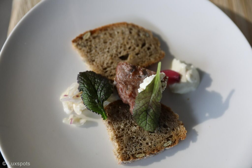 Ein Teller mit zwei Scheiben Brot, einer kleinen Portion Fleisch, garniert mit einem Blatt, und einem Klecks Sahne und Kohl.