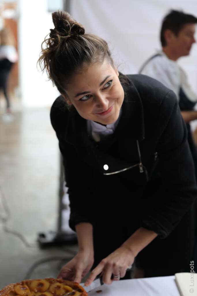 Eine junge Frau, die mit einem leichten Lächeln einen Kuchen schneidet und eine dunkle Jacke trägt.