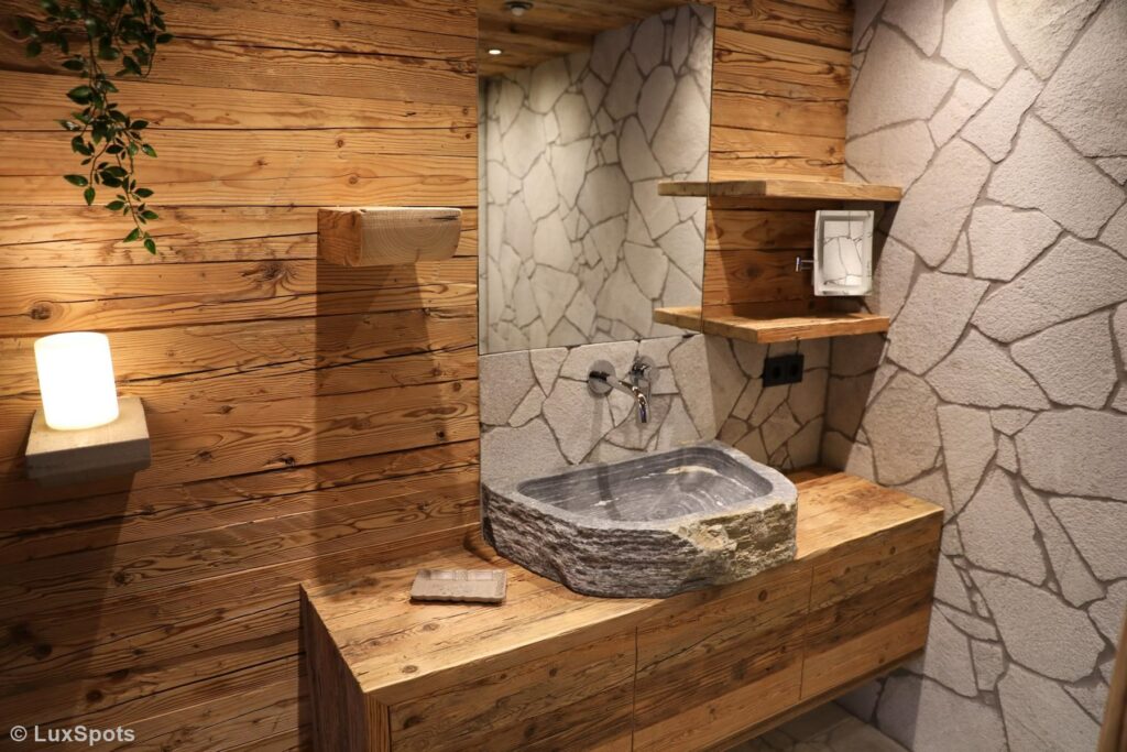 Rustikales Badezimmer mit Steinwaschbecken, Holzarbeitsplatte, Holzvertäfelung, Wänden mit Steinstruktur, Regalen und einer Wandleuchte.