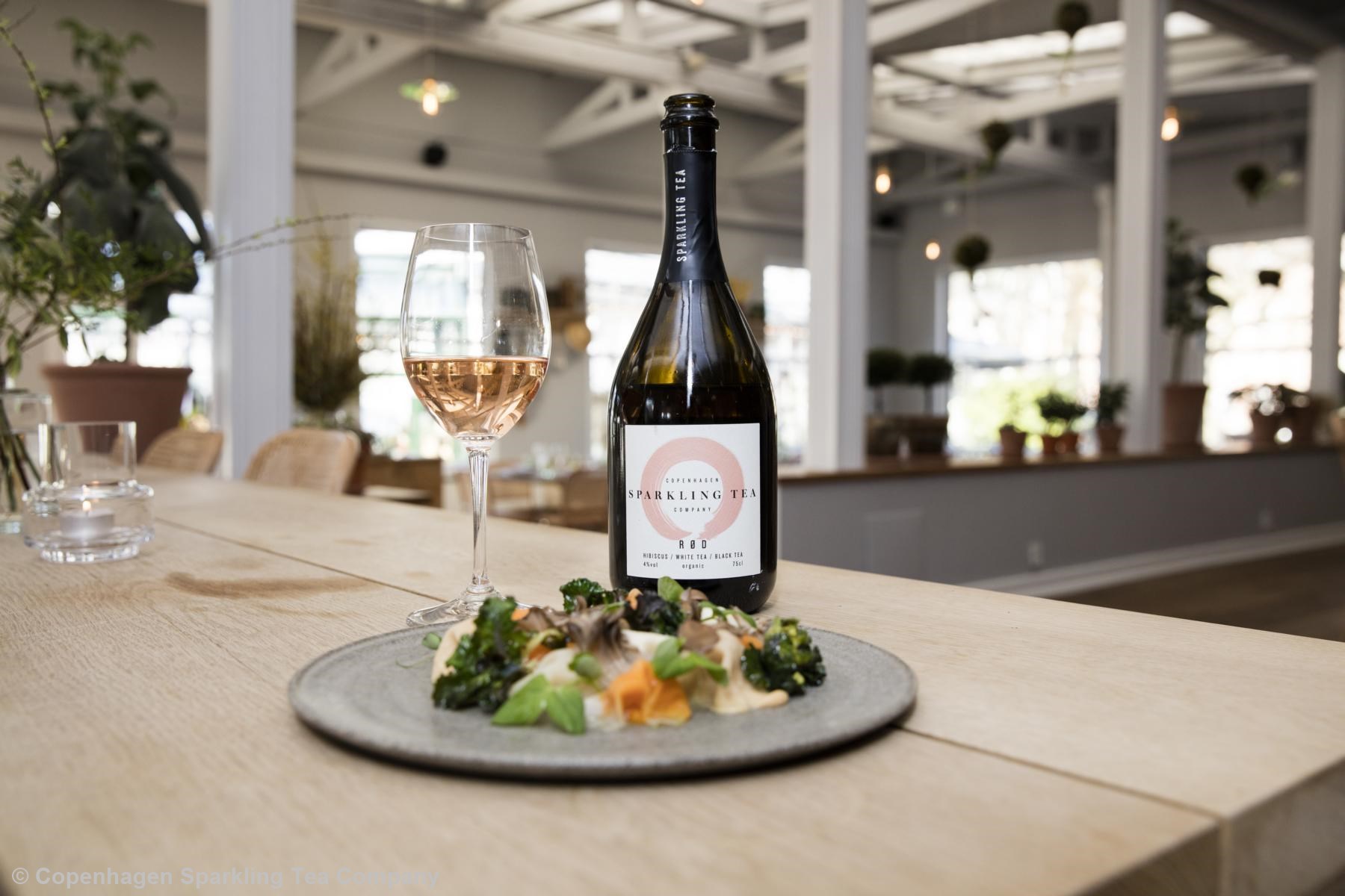 Eine Sektflasche steht mit einem rosefarbenen, alkoholfreien Sekt auf einem Tisch. Auf der Flasche steht "Sparkling Tea".