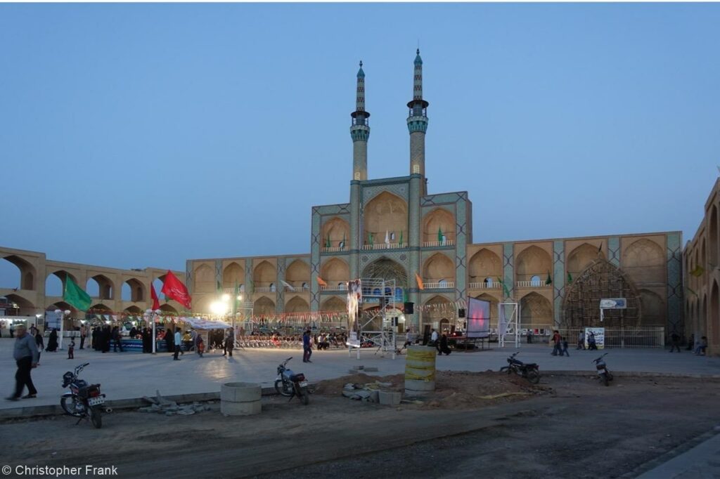 Ein altes, mehrstöckiges Gebäude mit zwei schmalen Türmen am Abend. Im Vordergrund sind eine Baustelle sowie Menschen auf einem Marktplatz zu erkennen.