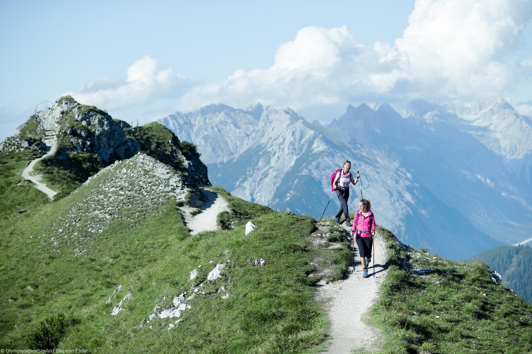 Zwei Frauen wandern mit Wanderstöcken und Rucksäcken auf dem Karwendel Gebirge in Tirol. Im Hintergrund sind weitere Berge udn Wolkenformationen zu sehen.