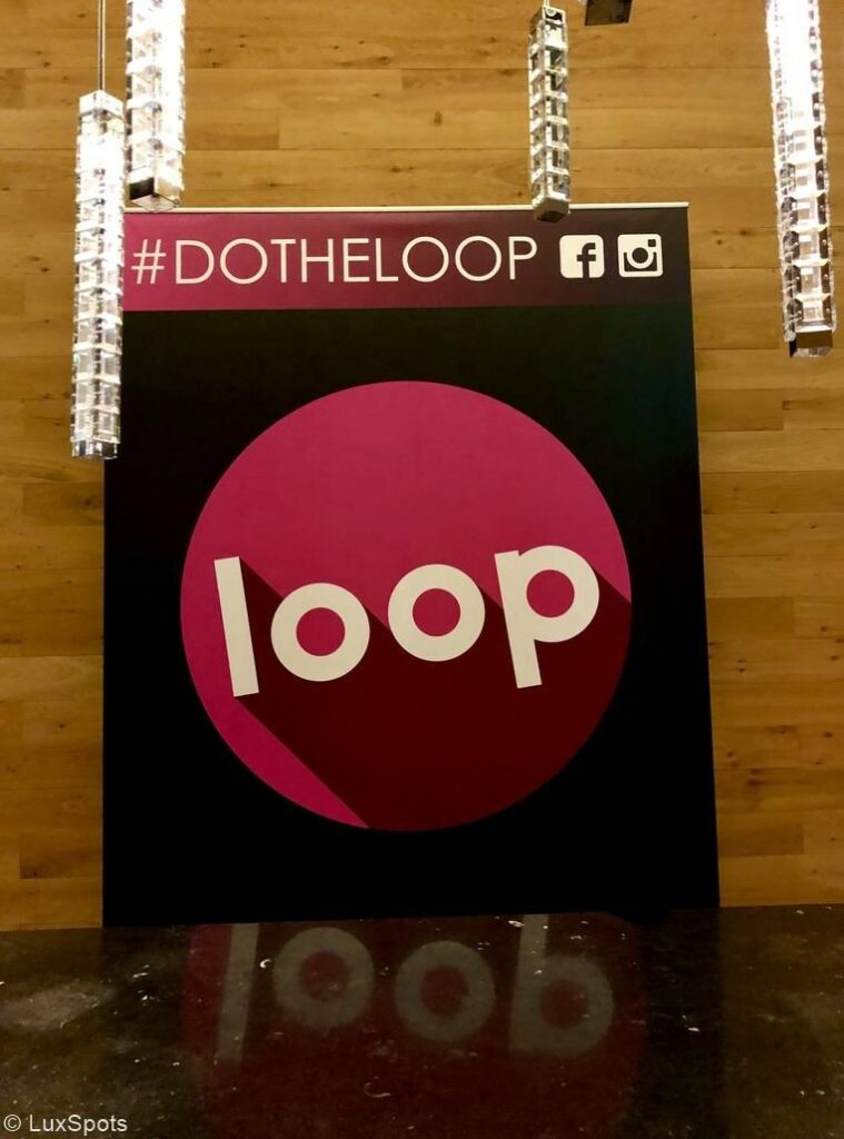 Schild mit „Loop“ in großen Buchstaben, „#DOTHELOOP“ darüber und Social-Media-Symbolen für Facebook und Instagram, vor einem holzgetäfelten Hintergrund mit Hängelampen.
