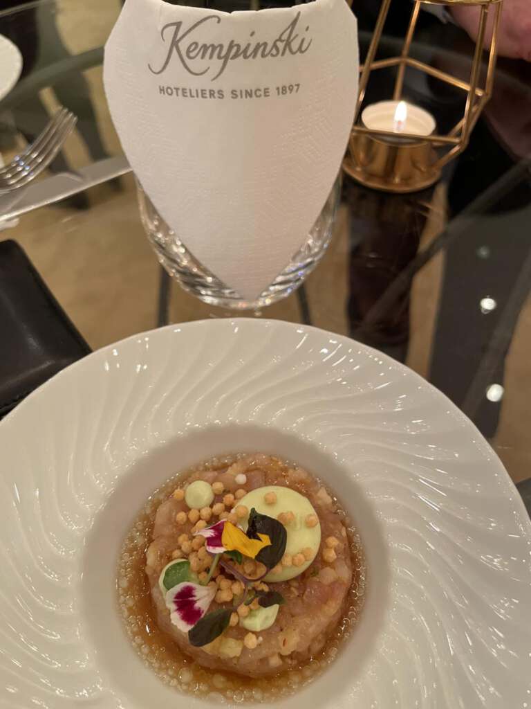 Ein Teller mit Fischtartar steht auf einem Glastisch. Dahinter ist eine Kempinski-Serviette im Glas sowie ein Teelicht zu sehen.