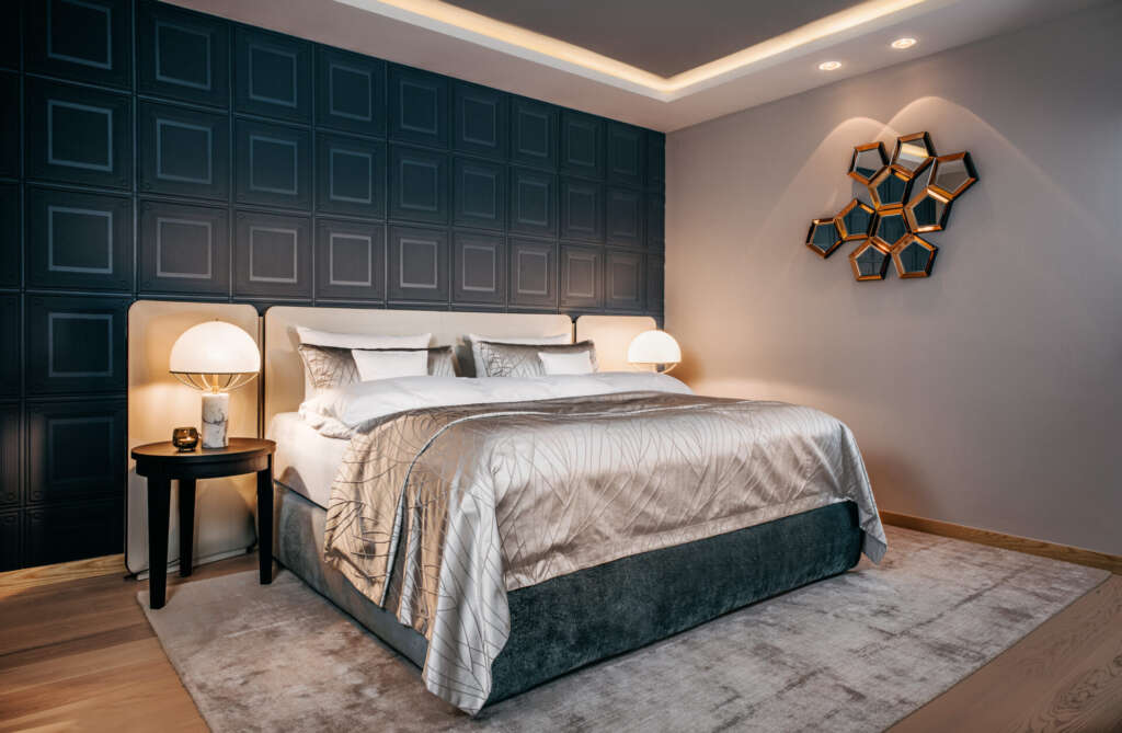 Modernes Schlafzimmer mit großem Bett, strukturierter dunkler Akzentwand, geometrischem Spiegel und zwei Beistelltischen mit Lampen.