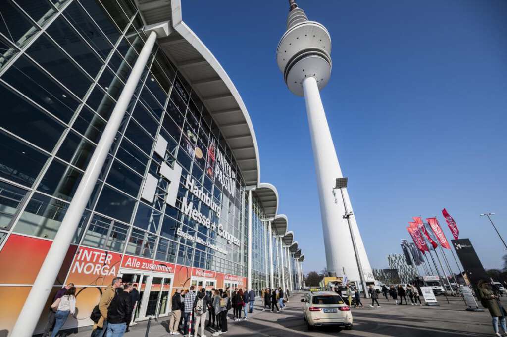 Menschen gehen vor dem Haupteingang der Messe Hamburg spazieren, in der Nähe thront der Heinrich-Hertz-Fernsehturm, davor wehen bunte Fahnen.