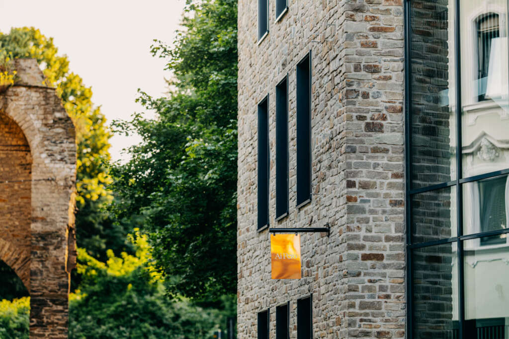 Steingebäude mit modernen Fenstern und einem kleinen gelb-orangefarbenen Schild an einem sonnigen Tag, das auf die Köstlichkeiten des Restaurants „Ai Pero“ hinweist. Bäume und ein teilweiser Blick auf eine Andernacher Bogenkonstruktion verstärken seinen Charme.