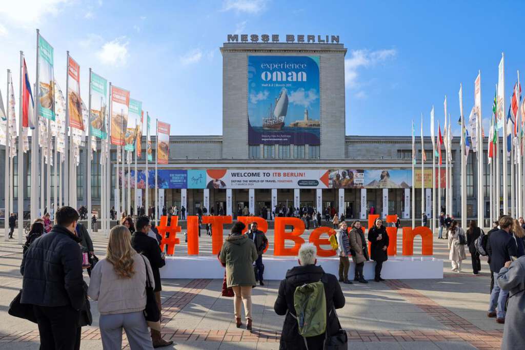 Menschenmenge vor der Messe Berlin mit „ITB Berlin“-Schild und internationalen Flaggen. Am Gebäude prangt ein großes Werbebanner mit der Aufschrift „Experience Oman“.