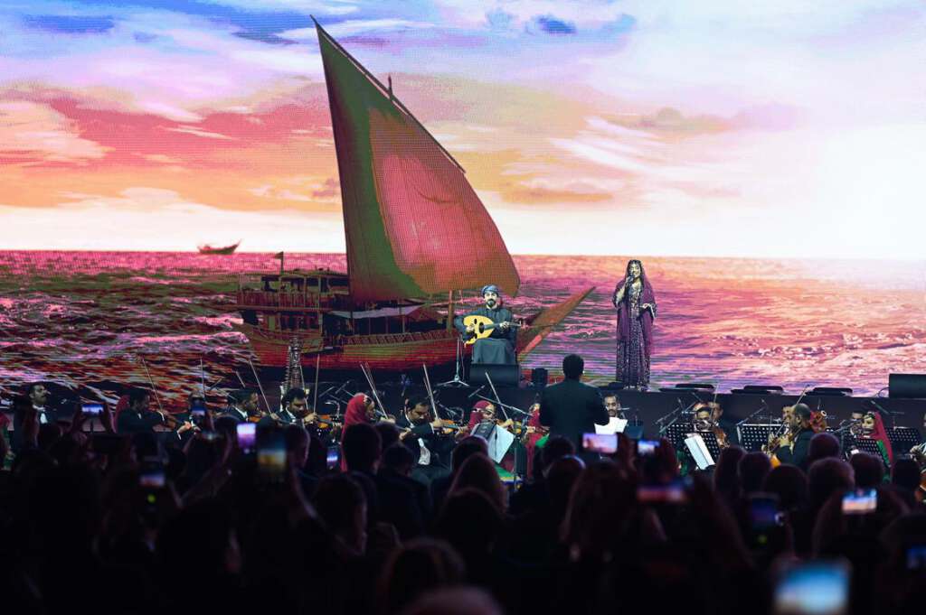 Ein Orchester spielt auf der Bühne vor der farbenfrohen Kulisse eines Segelboots auf dem Meer. Zuschauer machen mit ihren Handys Fotos.