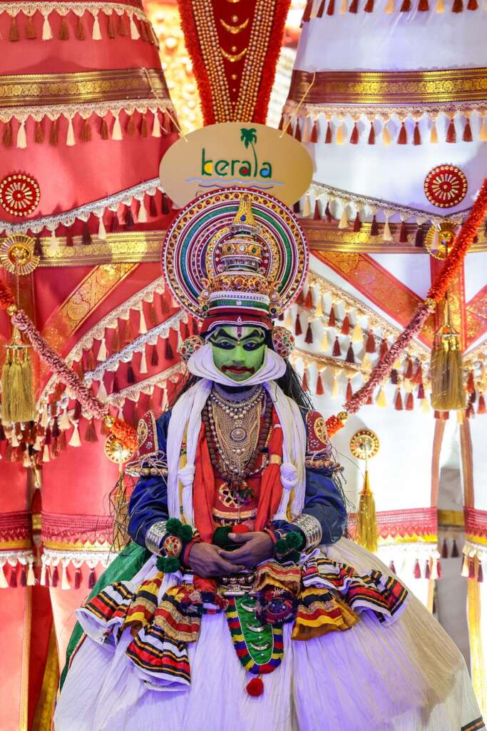 Eine Person im farbenfrohen traditionellen Kathakali-Kostüm und mit Make-up steht vor dekorierten Vorhängen, über denen ein „Kerala“-Schild steht.