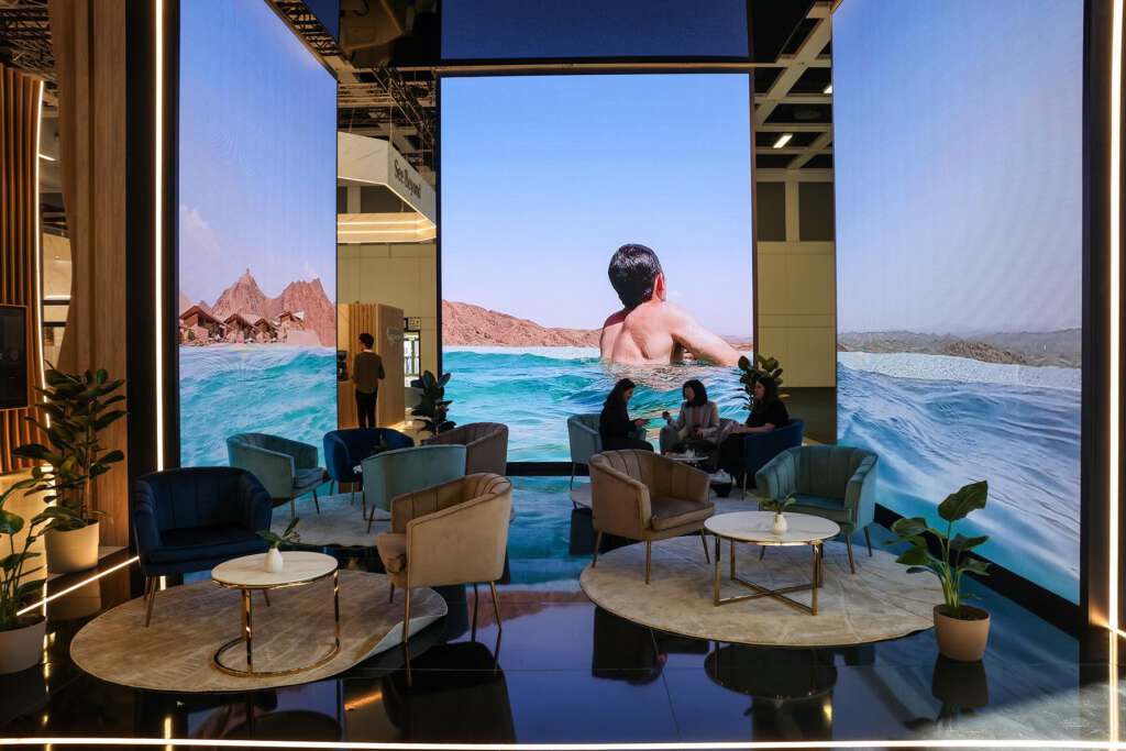 Ein moderner Loungebereich mit Sitzgelegenheiten und Tischen, umgeben von großen Bildschirmen, auf denen ein Mann zu sehen ist, der in einem blauen Ozean mit Bergen im Hintergrund schwimmt.