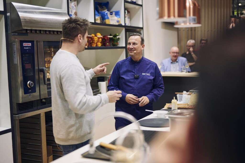 Zwei Männer unterhalten sich in einer modernen Küche. Einer trägt eine lila Kochjacke, der andere einen legeren Pullover. Auf der Arbeitsfläche sind Kochzutaten zu sehen.