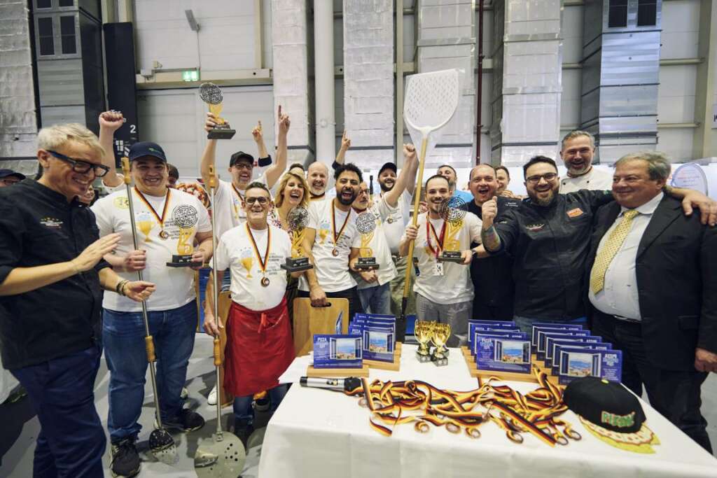 Eine Gruppe von Menschen feiert eine Veranstaltung und hält Trophäen und Medaillen in den Händen. Pizzaschaufeln sind zu sehen und auf einem Tisch im Vordergrund liegen Auszeichnungen.