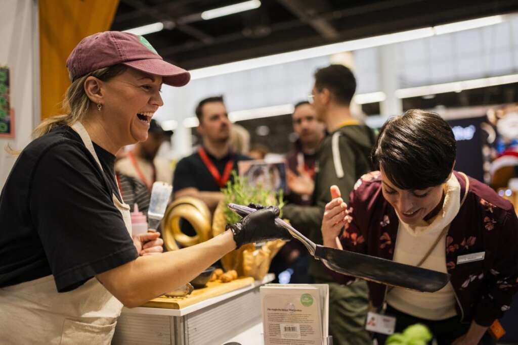 Eine Frau mit Schürze und Mütze serviert einem lächelnden Kunden auf einem geschäftigen Indoor-Markt oder bei einer Veranstaltung Essen.