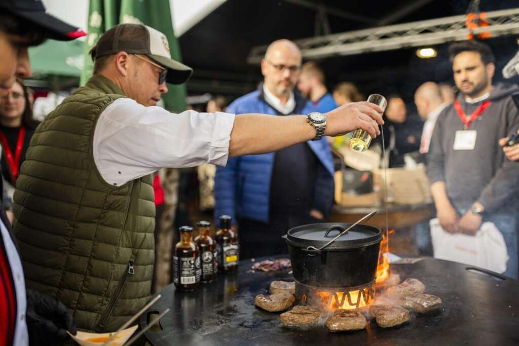 Ein Mann mit Mütze und Weste gießt Öl in einen Topf auf einem Außengrill, umgeben von Zuschauern. Auf dem Grill liegen Bierflaschen und Burger-Patties.