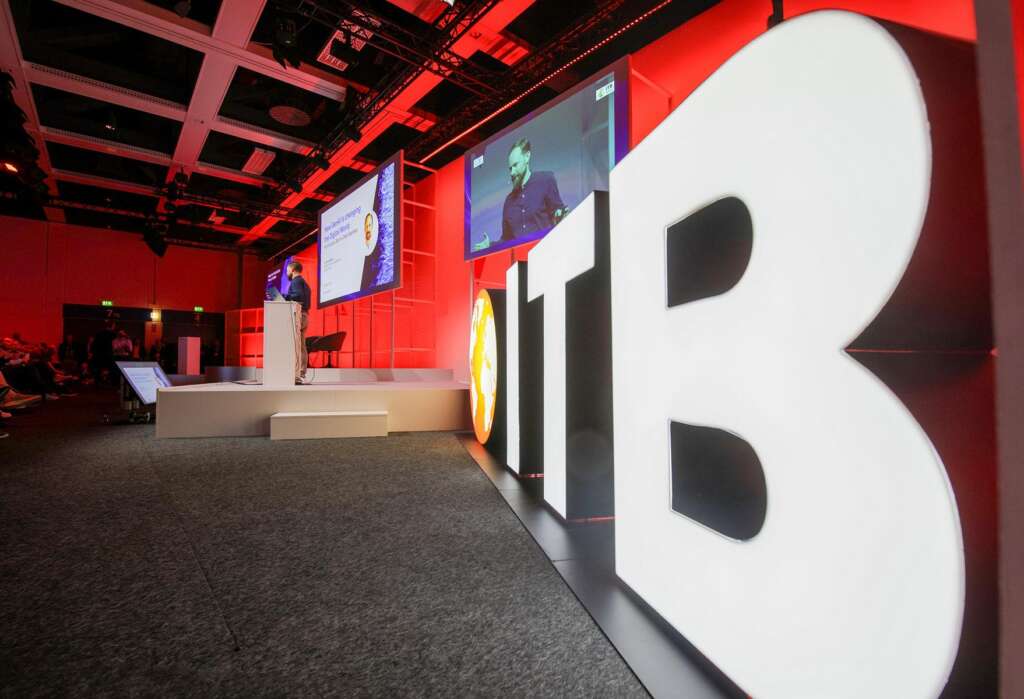In einem schwach beleuchteten Auditorium fesselt ein Redner das Publikum bei der ITB Berlin. Er steht neben einem großen Leuchtschild mit der Aufschrift „ITB“.