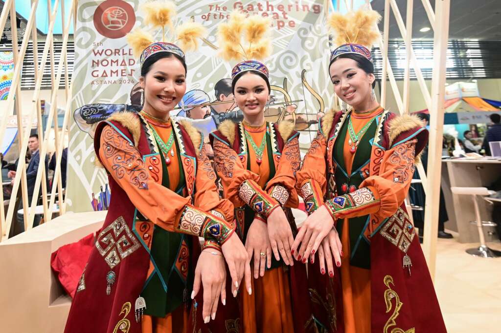 Drei Frauen in traditioneller Kleidung posieren mit ausgestreckten Händen an einem Veranstaltungsstand der World Nomad Games und schaffen eine lebendige Szene.