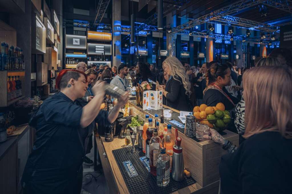 Eine belebte Barszene auf der Gastro Vision in Hamburg, mit einer Barkeeperin, die einen Cocktail-Shaker schüttelt, umgeben von Gästen und einer Auslage mit Obst und Getränkeflaschen.