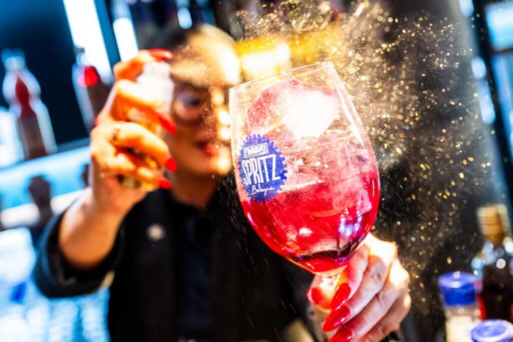 Eine Cocktailmixerin sprüht auf der Gastro Vision in Hamburg einen roten Cocktail an, der in einem mit dem „Spritz“-Logo verzierten Glas ist und erzeugt so einen zarten Sprühnebel.