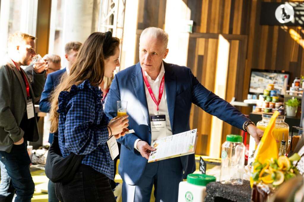 Auf der geschäftigen Gastro Vision-Fachmesse in Hamburg unterhalten sich zwei Personen. Eine Besucherin hält ein Getränk in der Hand, der Aussteller eine Angebotsliste vor einer Speise- und Getränkepräsentation.