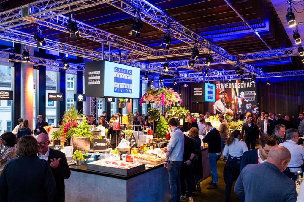 Menschen treffen sich in einem großen Innenraum des Hotels Empire Riverside zur Gastro Vision in Hamburg, mit hellen Lichtern und Blumendekorationen. Der Veranstaltungsort verfügt über mehrere Ausstellungstische und Deckenschilder, die verschiedene Aussteller hervorheben.