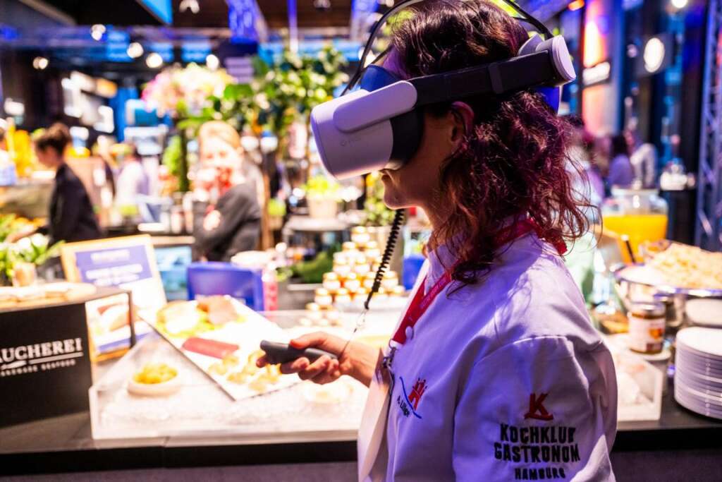 Eine Frau mit einem Virtual-Reality-Headset und einem Controller in der Hand steht in einem belebten Innenbereich der Gastro Vision, umgeben von verschiedenen Ausstellungsstücken und verlockenden Kulinarik-Angeboten.