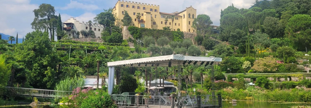 Ein See mit Seerosen und einer hierauf liegenden, überdachten Terrasse mit Steg zum Ufer. Im Hintergrund ist eine gelbfarbenes Gebäude mit Nachbargebäuden auf einem Hügel zu sehen.