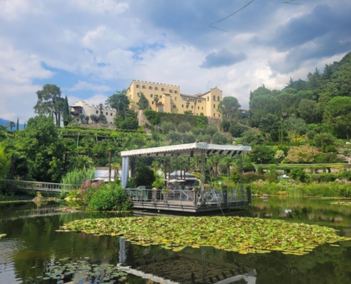 Ein See mit Seerosen und einer hierauf liegenden, überdachten Terrasse mit Steg zum Ufer. Im Hintergrund ist eine gelbfarbenes Gebäude mit Nachbargebäuden auf einem Hügel zu sehen.