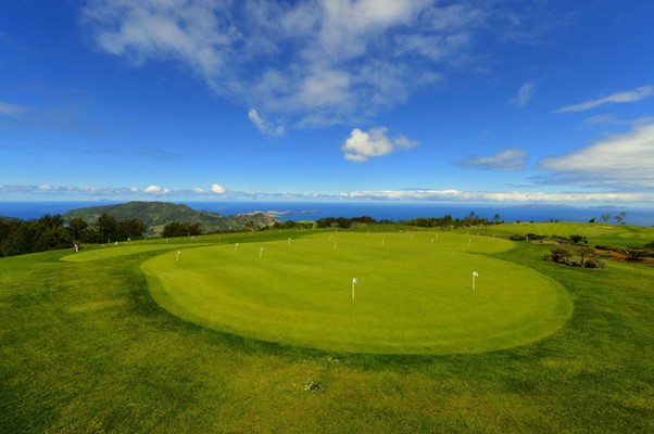Ein faszinierender Golfplatz auf der Insel Madeira, mit großem Übungsgrün und mehreren Flaggen, die die Übungslöcher markieren. Es ist von sattgrünen Hügeln umgeben und bietet unter einem leicht bewölkten Himmel Ausblick auf das Meer.
