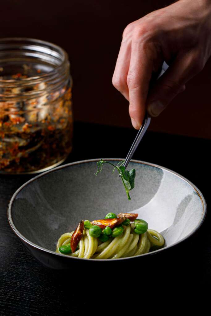 Eine Nahaufnahme eines Gerichts, das mit frischen Kräutern mithilfe einer Pinzette garniert wird. Das Gericht besteht aus Pasta, leuchtend grünen Erbsen und knusprigen Fleischstücken, vor einem dunklen Hintergrund, mit einem Gewürzglas im Hintergrund.