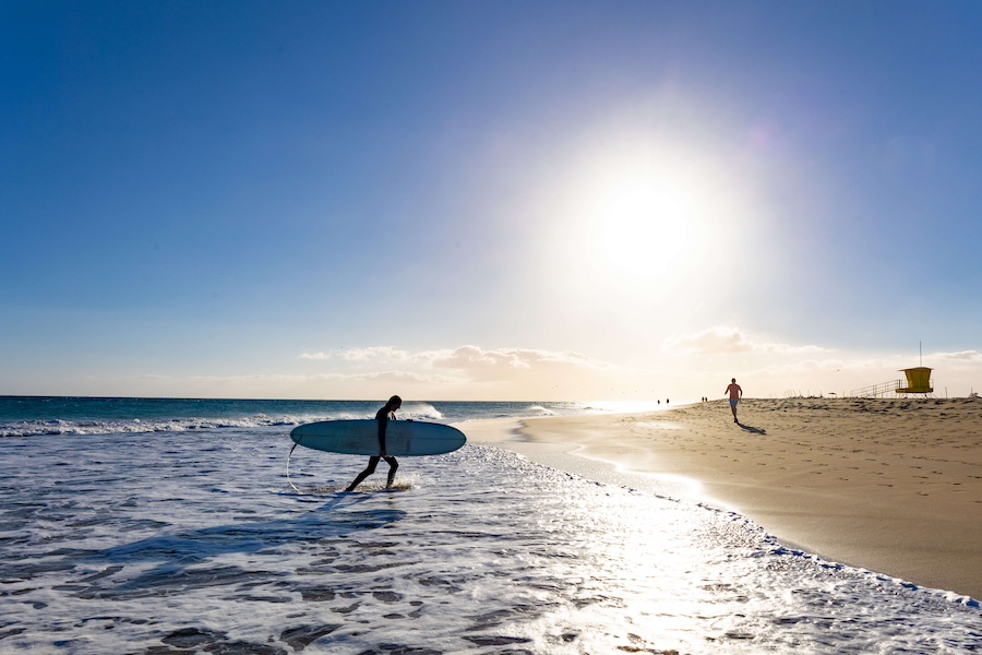 Ein Surfer geht mit einem Surfbrett durch flaches Wasser am Strand von Morro Jable auf Fuerteventura. Die Sonne steht tief am Himmel, und das Bild zeigt einen klaren blauen Himmel sowie die weite Sandküste.