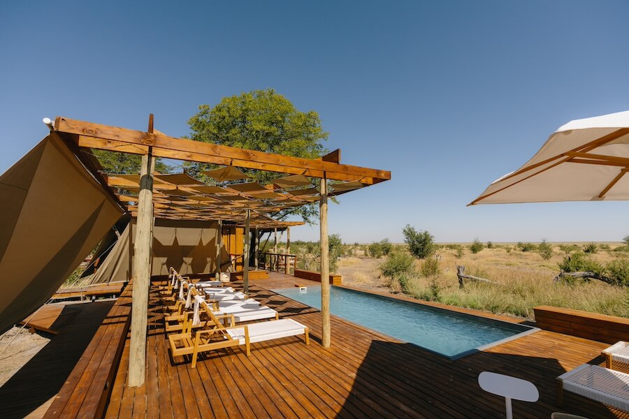 Eine luxuriöse Safari-Lodge mit einem hölzernen Sonnendeck und einem schlanken Pool im Vordergrund. Der Pool ist von bequemen Liegestühlen umgeben, und darüber spenden große Sonnenschirme und eine Pergola aus Holz Schatten. Die umgebende Landschaft ist eine weite, offene Steppe unter einem klaren blauen Himmel.