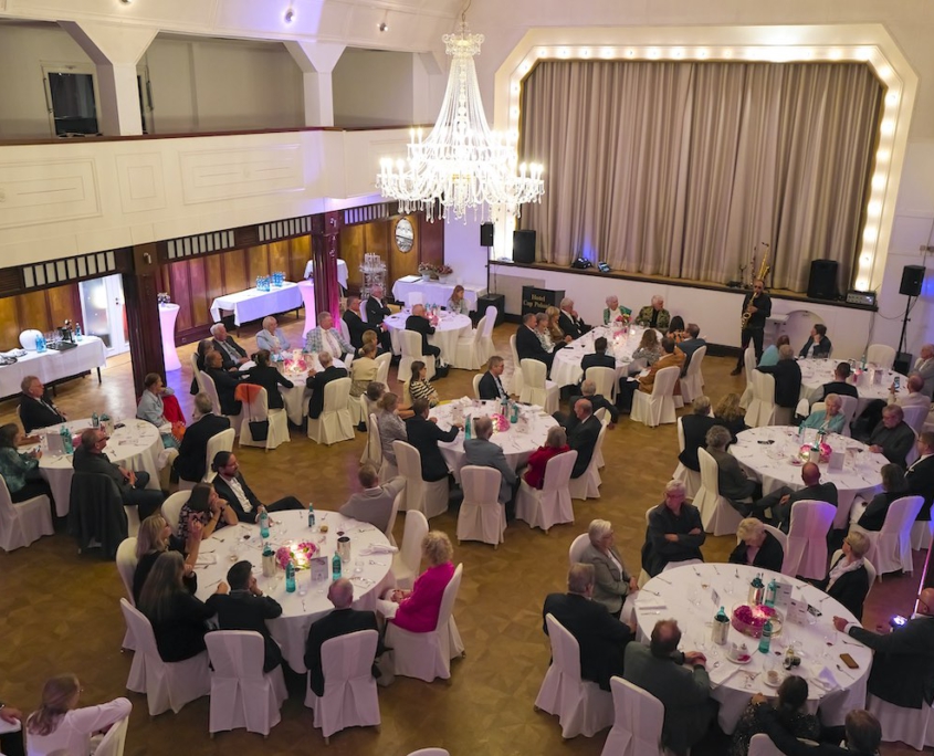 Blick von oben in einen Saal eines Hotels in Pinneberg bei Hamburg. Mehrere Gäste sitzen jeweils um einen großen, runden Tisch und unterhalten sich, während hinten rechts vor einer Bühne ein Saxophonist Musik spielt.