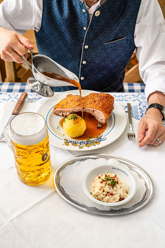 Ein Mann gießt Sauße auf sein Spanferkelbraten mit Kloß. Daneben steht ein großes Bier und ein Krautsalat als Beilage.