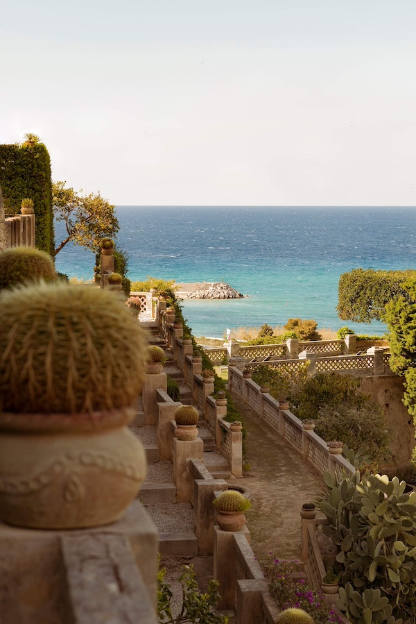 Terrassengarten mit Kakteentöpfen und Blick auf das klare blaue Mittelmeer unter einem hellblauen Himmel. Steingeländer führen hinunter zum Wasser und erinnern an einen ruhigen Zufluchtsort in Kalabrien.