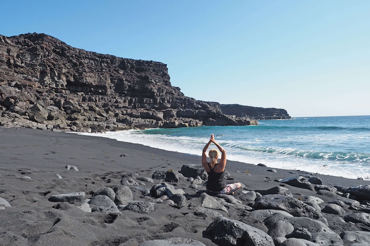 Eine Person praktiziert Yoga auf einem schwarzen Vulkanstrand, umgeben von großen Felsen. Im Hintergrund sind das Meer und eine felsige Klippe zu sehen. Die Person sitzt im Lotussitz und streckt die Arme nach oben in einer Meditationspose.