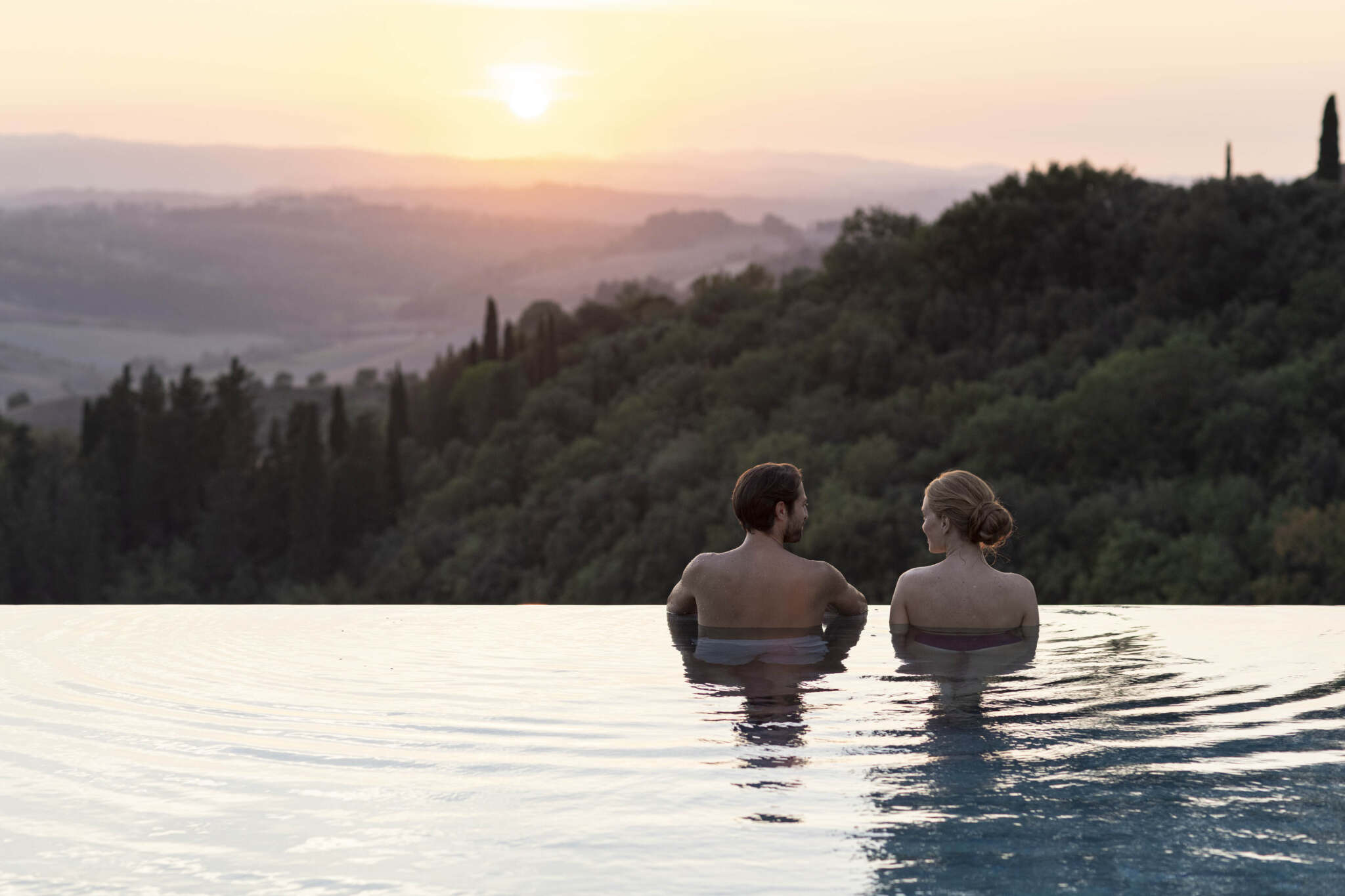Zwei Personen stehen im Wasser am Rande eines Infinity-Pools und genießen den Blick auf Hügel und Bäume bei Sonnenuntergang.