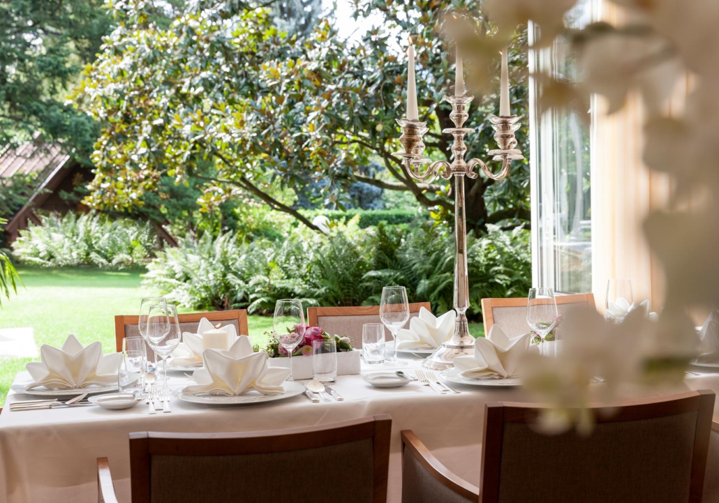 Ein langer Tisch steht schön und edel gedeckt sowie mit einem silberfarbenen Kerzenhalter im Mittelpunkt dieses Bildes. Im Hintergrund ist ein großer Garten zu sehen.