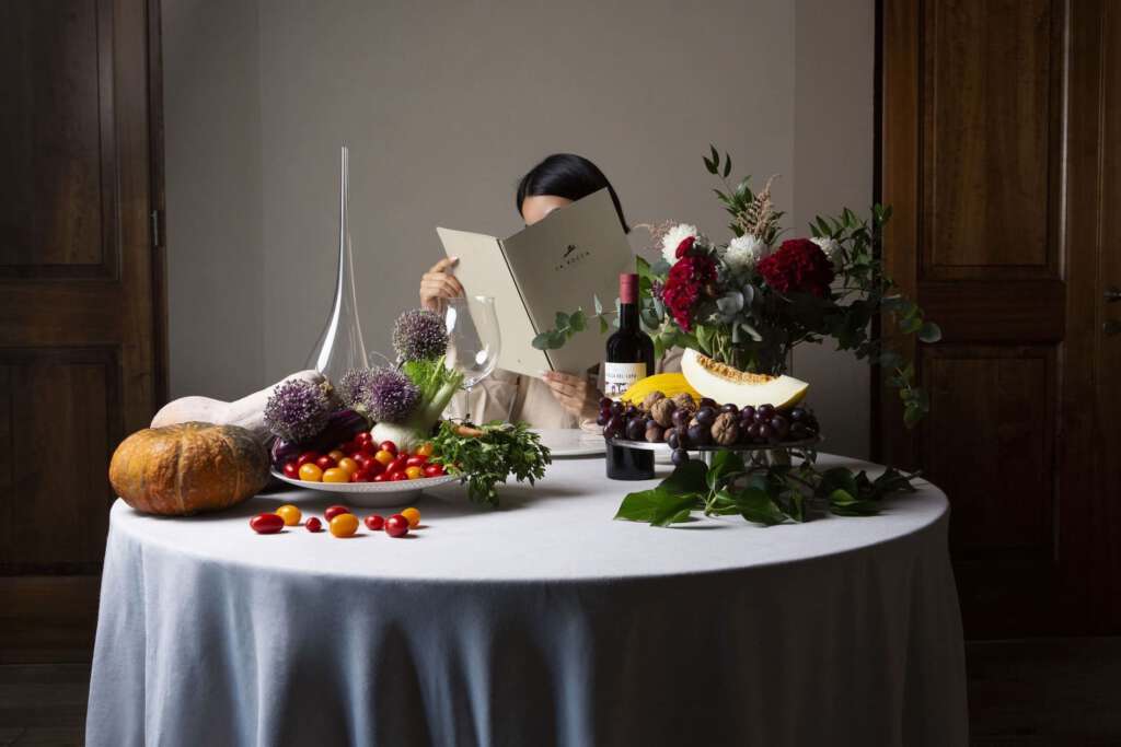 Eine Person hinter einer Speisekarte sitzt an einem Tisch mit einer Auswahl an Obst, Gemüse, Weinflaschen und Blumenarrangements.